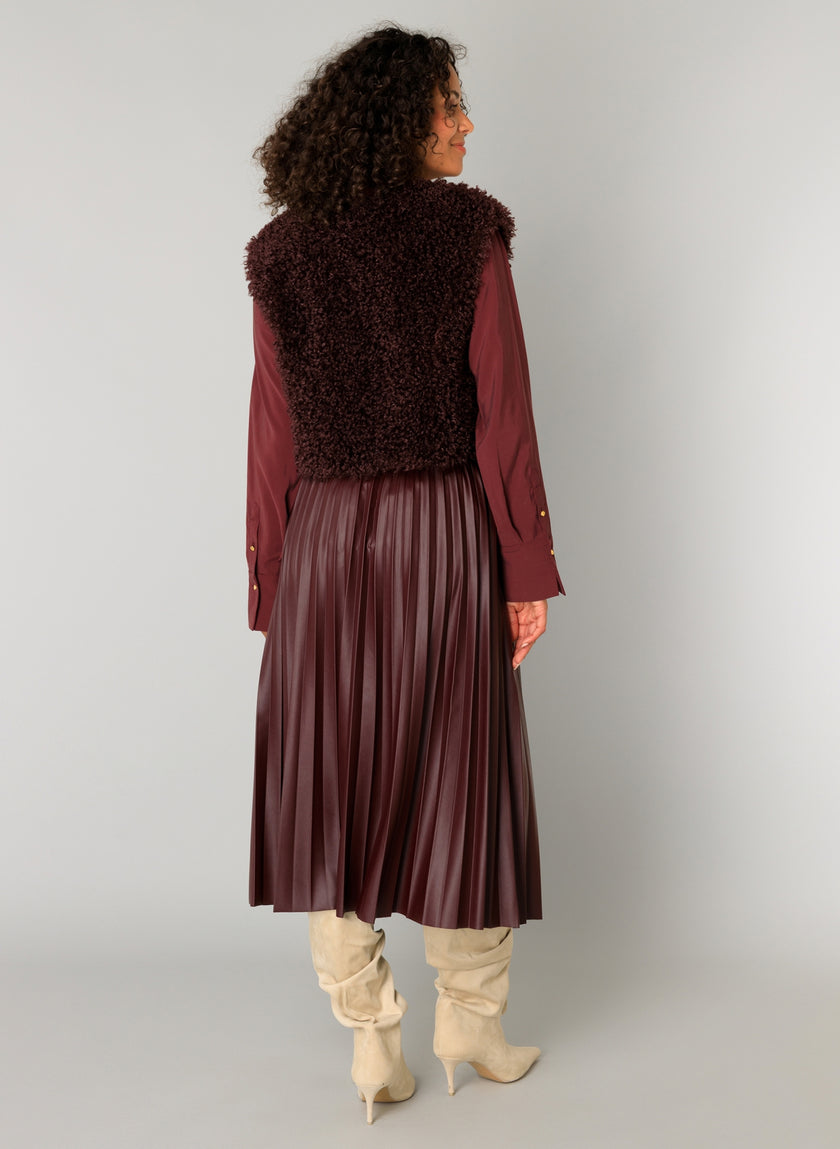 Een vrouw kijkt de andere kant op en draagt de Caroline - Donker bordeaux geplooide midirok met een bordeauxrode blouse met lange mouwen, een zacht laagjesvest van korte vezels en slouchy crèmekleurige kniehoge laarzen.