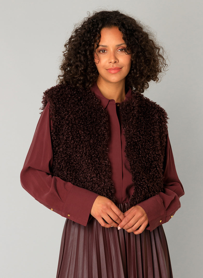 Een vrouw met krullend haar draagt de Caroline - Donker Bordeaux kastanjebruine blouse en een zacht, kortharig donkerbruin structuurvest, staande tegen een lichtgrijze achtergrond. Ze kijkt naar de camera met een neutrale uitdrukking en haar handen in elkaar geklemd.