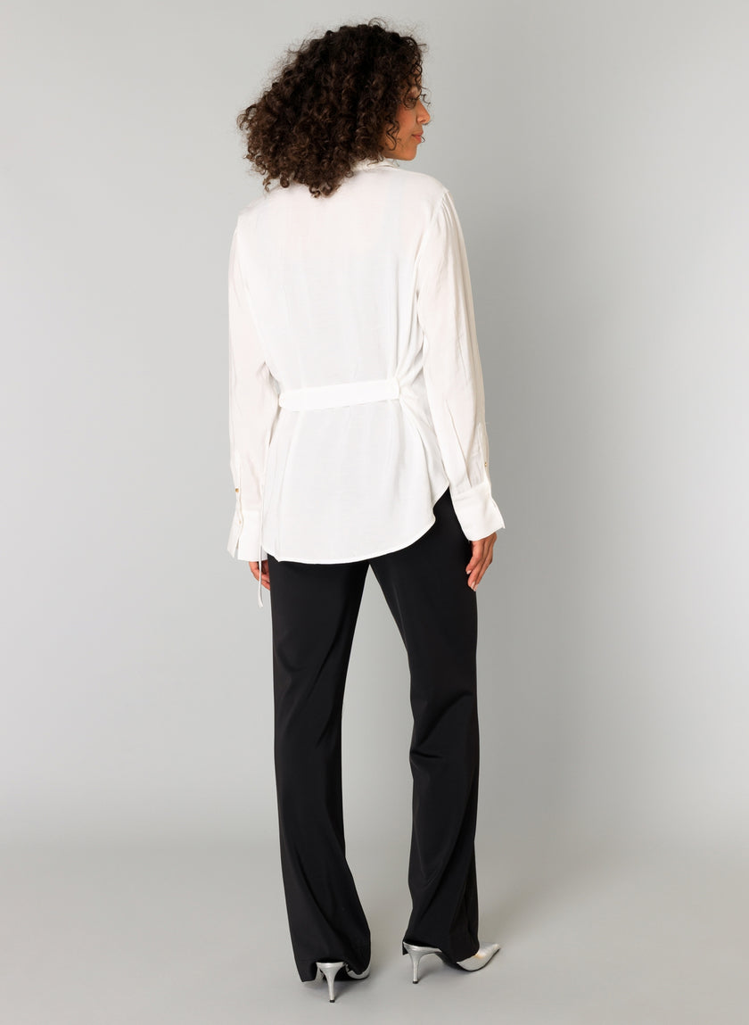 Een vrouw met krullend haar staat van zich af te kijken en draagt de Clara - Off White viscosemix damesblouse met een strikceintuur, een zwarte broek en witte hakken met spitse tenen tegen een effen lichtgrijze achtergrond.