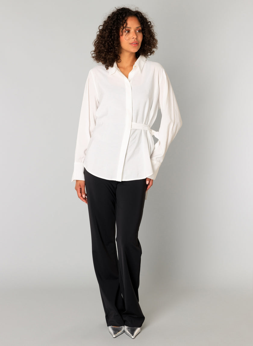 Een vrouw met krullend haar draagt de Clara - Off White damesblouse met lange mouwen en een strikceintuur, een zwarte broek en zilveren puntschoenen, tegen een effen lichtgrijze achtergrond.