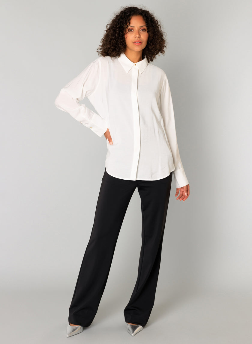 Een vrouw met krullend haar staat tegen een lichtgrijze achtergrond en draagt de Clara - Off White blouse, zwarte broek en zilveren hakken met spitse teen. Ze poseert zelfverzekerd met één hand op haar heup en de andere naast haar zijde.