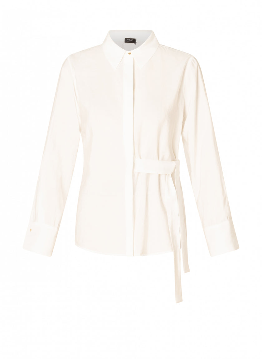 De Clara - Off White blouse is een witte viscosemix top met lange mouwen, een spitse kraag, verborgen knoopsluiting, brede manchetten en een opvallende ceintuur in de taille, tegen een effen witte achtergrond.