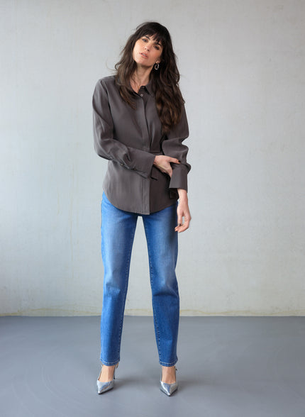 Een vrouw met lang donker haar staat binnen tegen een lichtgrijze muur. Ze draagt de Clara - Chocolate Brown blouse in zachte viscosemix, gecombineerd met een blauwe jeans en zilveren hakken met spitse tenen. Eén hand rust naast haar zijde, de andere raakt haar arm aan.