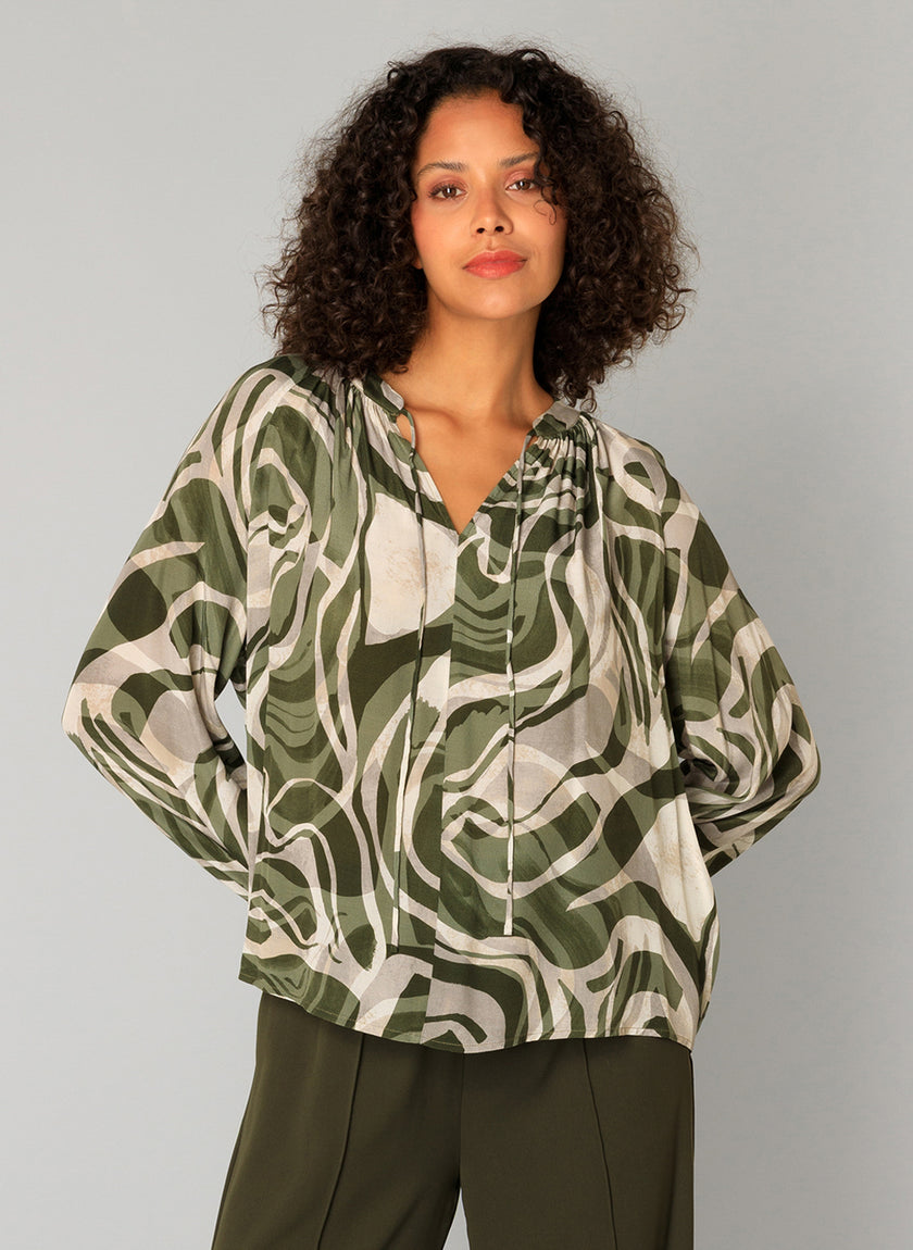 Een vrouw met krullend haar draagt de Carmen - Legergroene viscose blouse met een organische print, v-hals en wijde mouwen, gecombineerd met een bijpassende groene broek, tegen een effen lichtgrijze achtergrond.