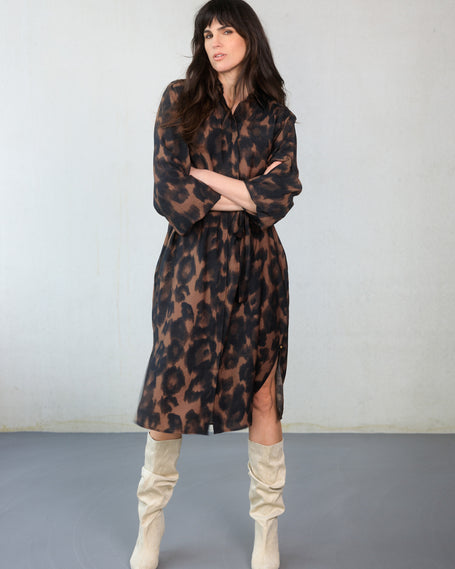 Een vrouw met lang donker haar poseert zelfverzekerd in de Charlene, een transparante blousejurk met dierenprint, lange mouwen en een strikceintuur, gecombineerd met slouchy, lichtgekleurde kniehoge laarzen tegen een effen, lichte achtergrond.