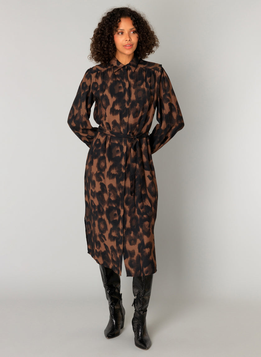 Een vrouw met krullend haar draagt de Charlene lange mouwen blousejurk in bruin-zwart animalprint met ceintuur en midi lengte, gecombineerd met zwarte knielaarzen. Ze poseert tegen een effen lichtgrijze achtergrond.