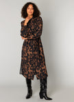 Een vrouw met krullend haar showt de Charlene blousejurk in bruine en zwarte dierenprint met midi lengte en lange mouwen, gestyled met zwarte kniehoge cowboylaarzen tegen een effen lichtgrijze achtergrond.