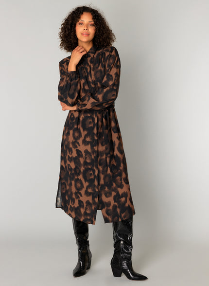 Een vrouw met krullend haar showt de Charlene blousejurk in bruine en zwarte dierenprint met midi lengte en lange mouwen, gestyled met zwarte kniehoge cowboylaarzen tegen een effen lichtgrijze achtergrond.