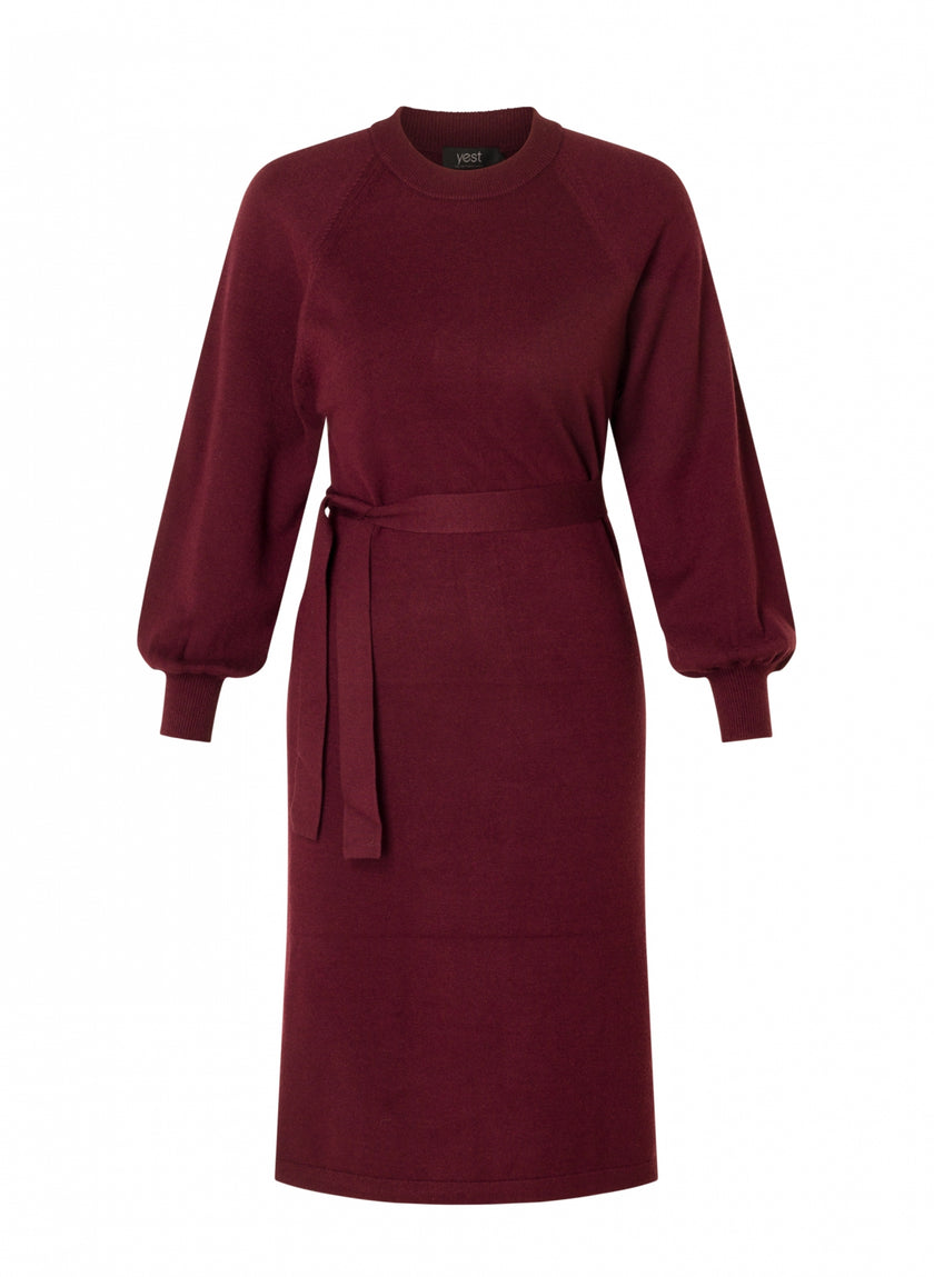 De Neva Essential is een kastanjebruine, knielange gebreide jurk met lange mouwen, een ronde halslijn en een ceintuur in de taille, tegen een effen witte achtergrond.