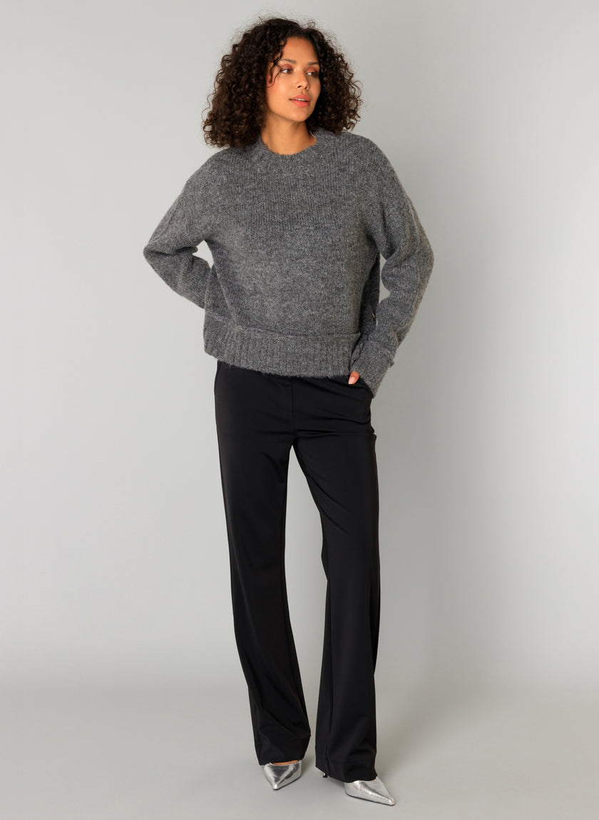 Een persoon met krullend haar draagt de Olieve Essential grofgebreide trui gemaakt van gerecyclede vezels en paart deze met een zwarte geklede broek en zilveren schoenen met spitse tenen. Hij staat tegen een effen lichtgrijze achtergrond en heeft één hand in zijn zak.