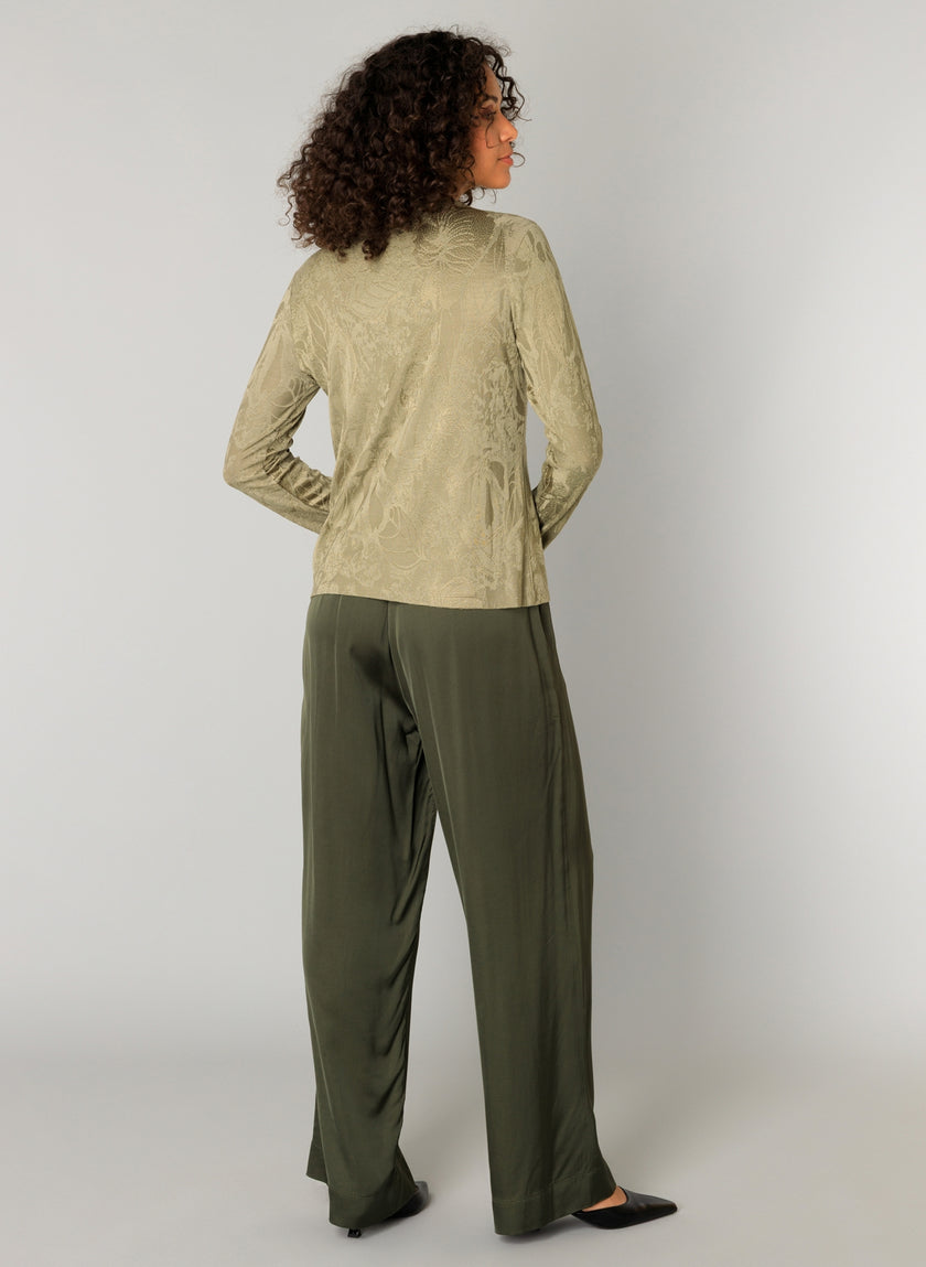 Een vrouw met krullend haar kijkt de andere kant op en draagt het Constança - Sage shirt en een donker olijfkleurige broek met wijde pijpen en zwarte puntschoenen tegen een effen lichtgrijze achtergrond.