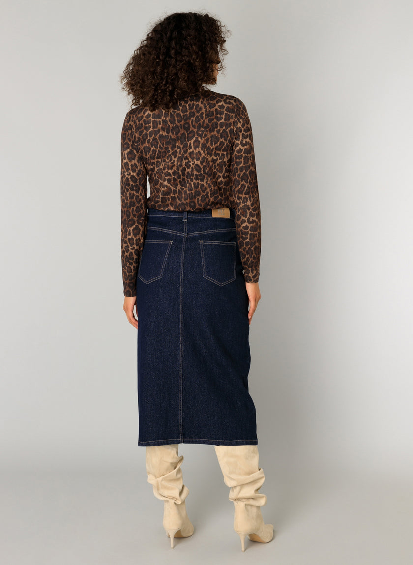 Een persoon met krullend haar staat van zich af te kijken en draagt de Omani Essential - Leopard top met lange mouwen, een donkerblauwe denim midi-rok en slouchy beige kniehoge laarzen tegen een effen lichte achtergrond.