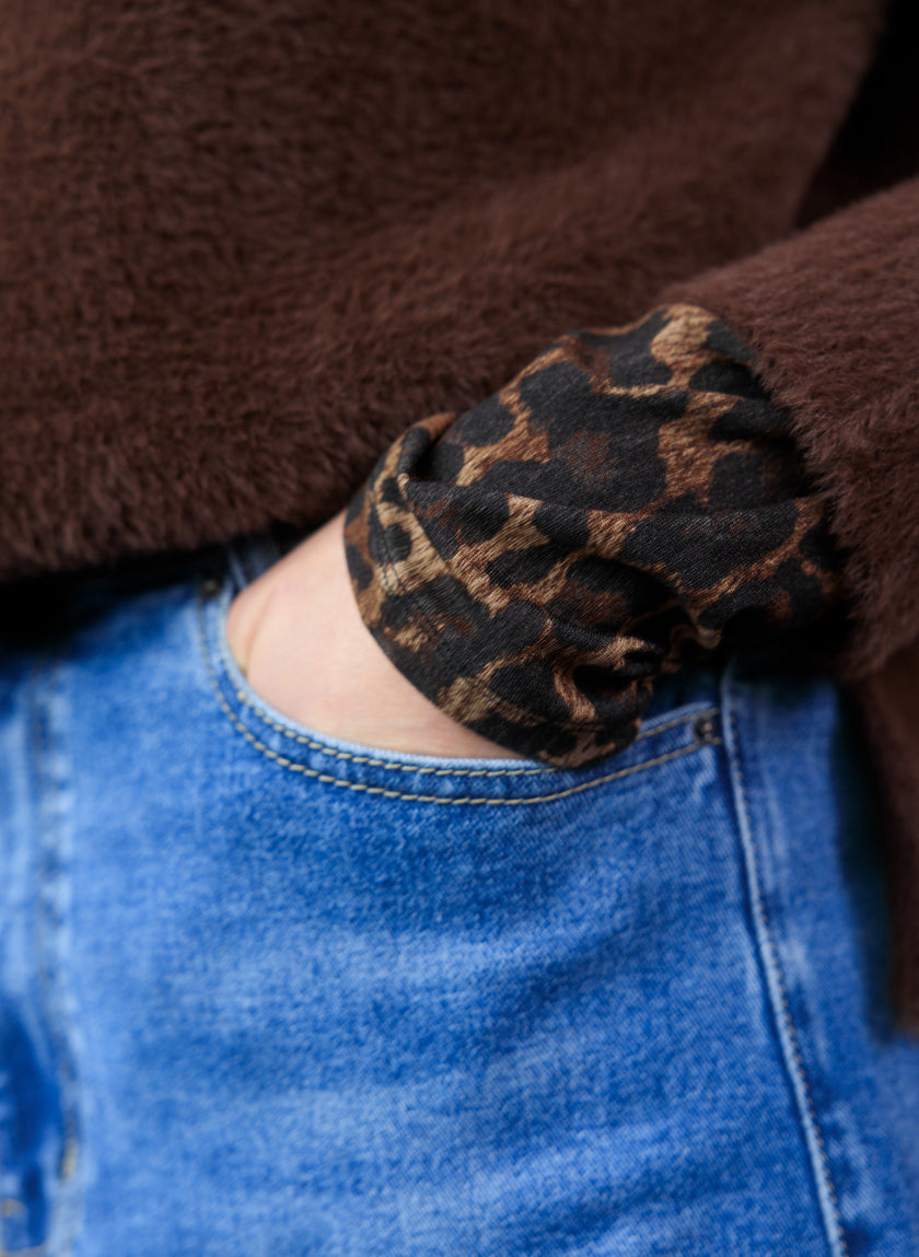 Een persoon met een blauwe spijkerbroek en de Omani Essential - Leopard bruine fuzzy sweater (stijl 0005856), met een manchet met luipaardprint, heeft zijn hand in de zak van zijn spijkerbroek gestoken.