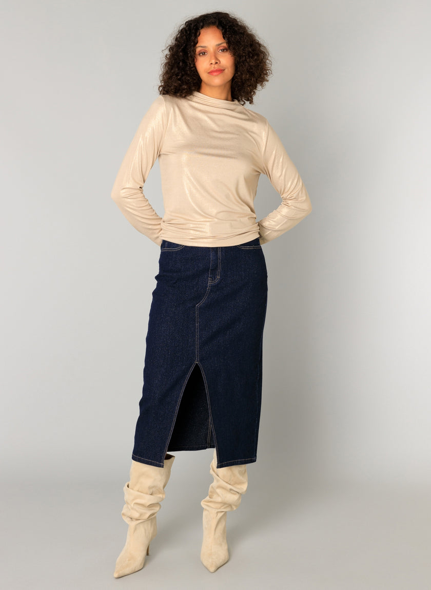Een persoon met krullend haar draagt de Candela - Sand slouchy kniehoge beige laarzen en combineert ze met een glanzende beige slim fit viscosemix longsleeve top en een donkerblauwe denim midi-rok met split, tegen een lichtgrijze achtergrond.