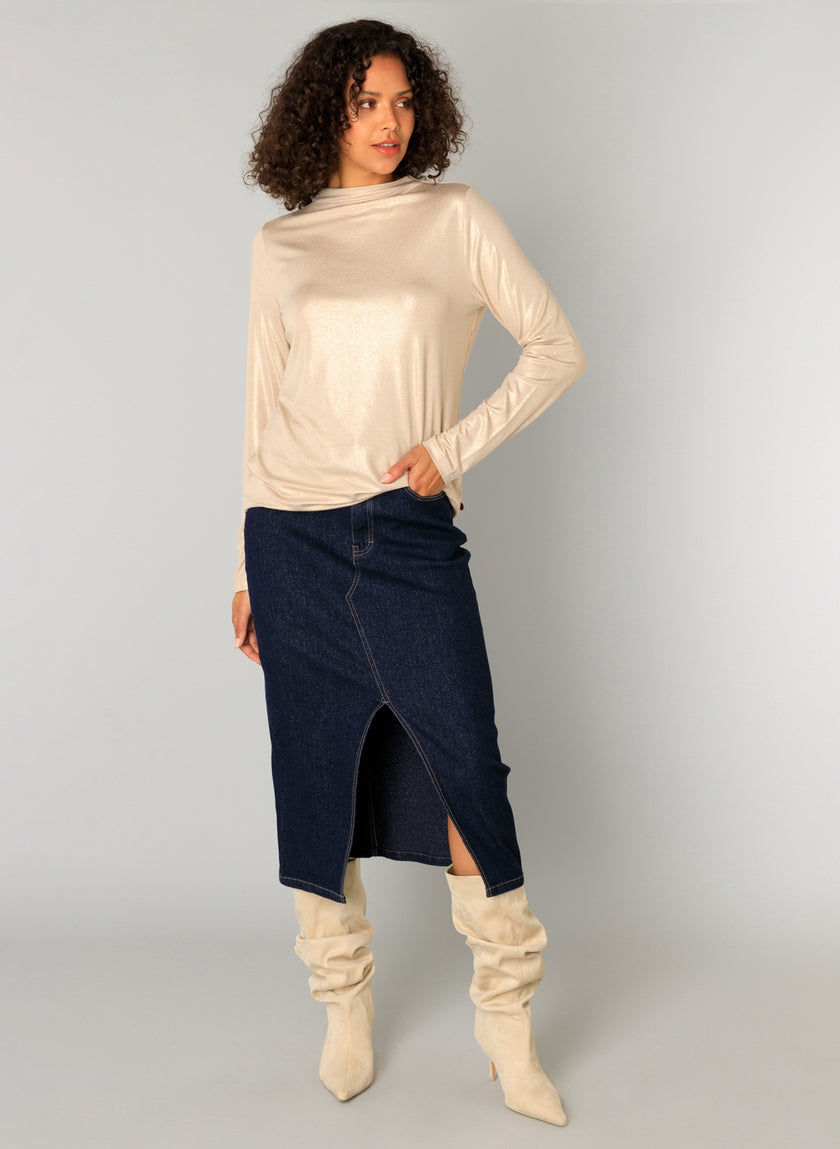 Een vrouw met krullend haar draagt een slim fit beige viscosemix top met lange mouwen, een donkerblauwe denim midi-rok met een split aan de voorkant en Candela - Sand slouchy kniehoge laarzen, staande tegen een effen lichtgrijze achtergrond.