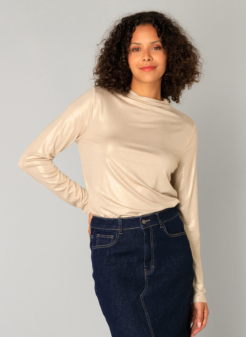 Een vrouw met krullend haar draagt de Candela - Sand glimmende viscosemix longsleeve top en een donkere denim rok, staande tegen een lichtgrijze achtergrond, zacht glimlachend met een hand op haar heup.