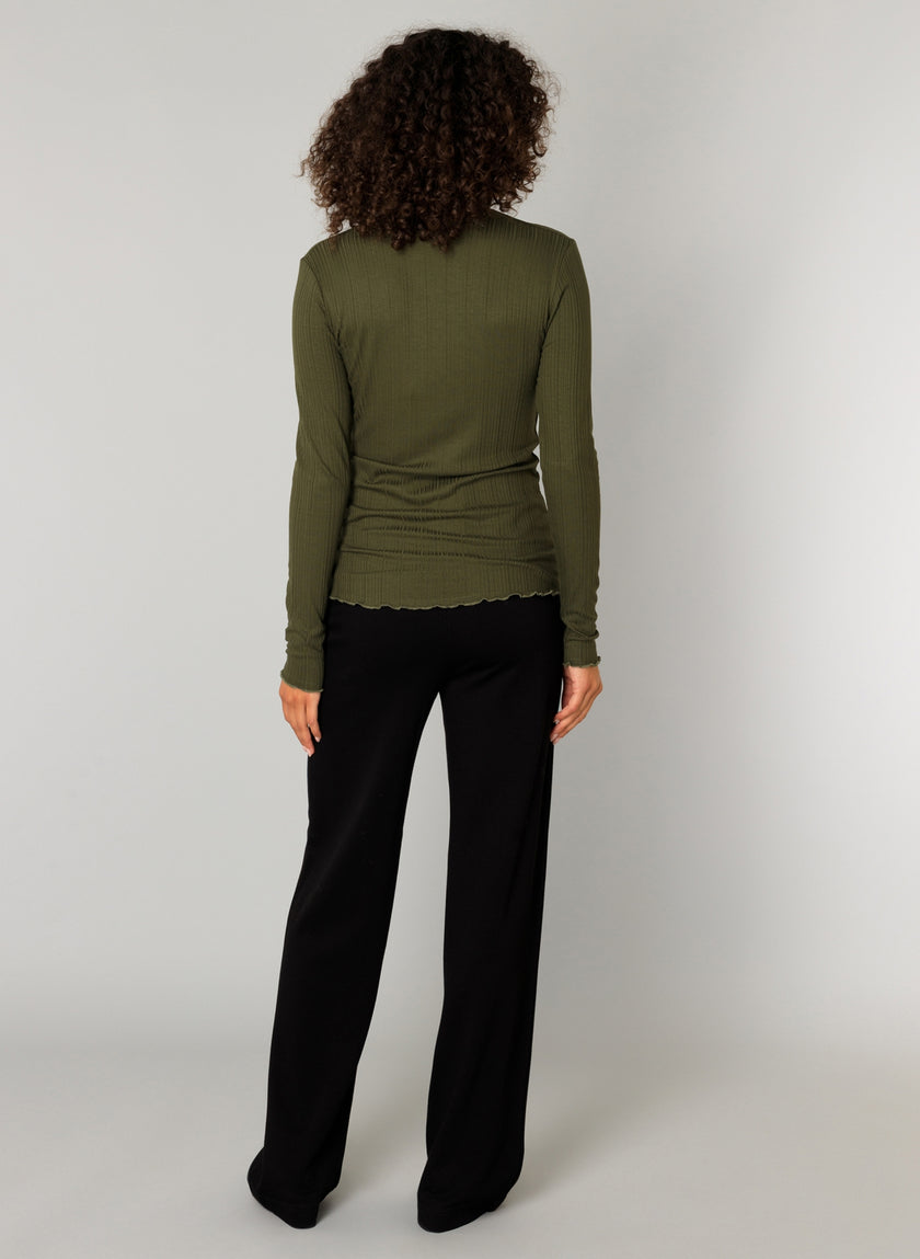 Een persoon met krullend haar staat van zich af te kijken, draagt een olijfgroene top met lange mouwen en de Paloma Essential zachte pantalon met elastische tailleband, beschikbaar in 4 kleuren, tegen een effen lichte achtergrond.