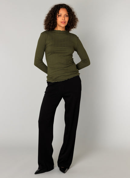 Een vrouw met krullend haar staat tegen een effen achtergrond en draagt de Paloma Essential in olijfgroen met een zachte elastische tailleband pantalon en zwarte puntschoenen. Ze kijkt naar voren, handen achter haar rug. Nu verkrijgbaar in 4 kleuren.