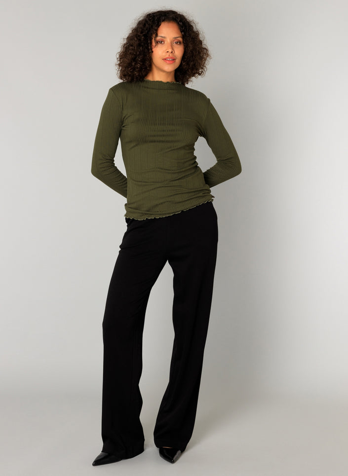 Een vrouw met krullend haar staat tegen een effen achtergrond en draagt de Paloma Essential in olijfgroen met een zachte elastische tailleband pantalon en zwarte puntschoenen. Ze kijkt naar voren, handen achter haar rug. Nu verkrijgbaar in 4 kleuren.