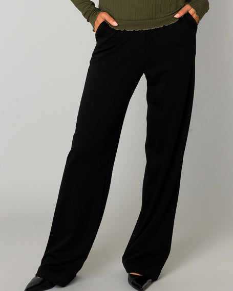 Een persoon staat tegen een effen lichtgrijze achtergrond en draagt de Paloma Essential zwarte zachte pantalon met elastische tailleband, een geribde olijfgroene top met lange mouwen en zwarte puntschoenen. De Paloma Essential is beschikbaar in 4 kleuren.