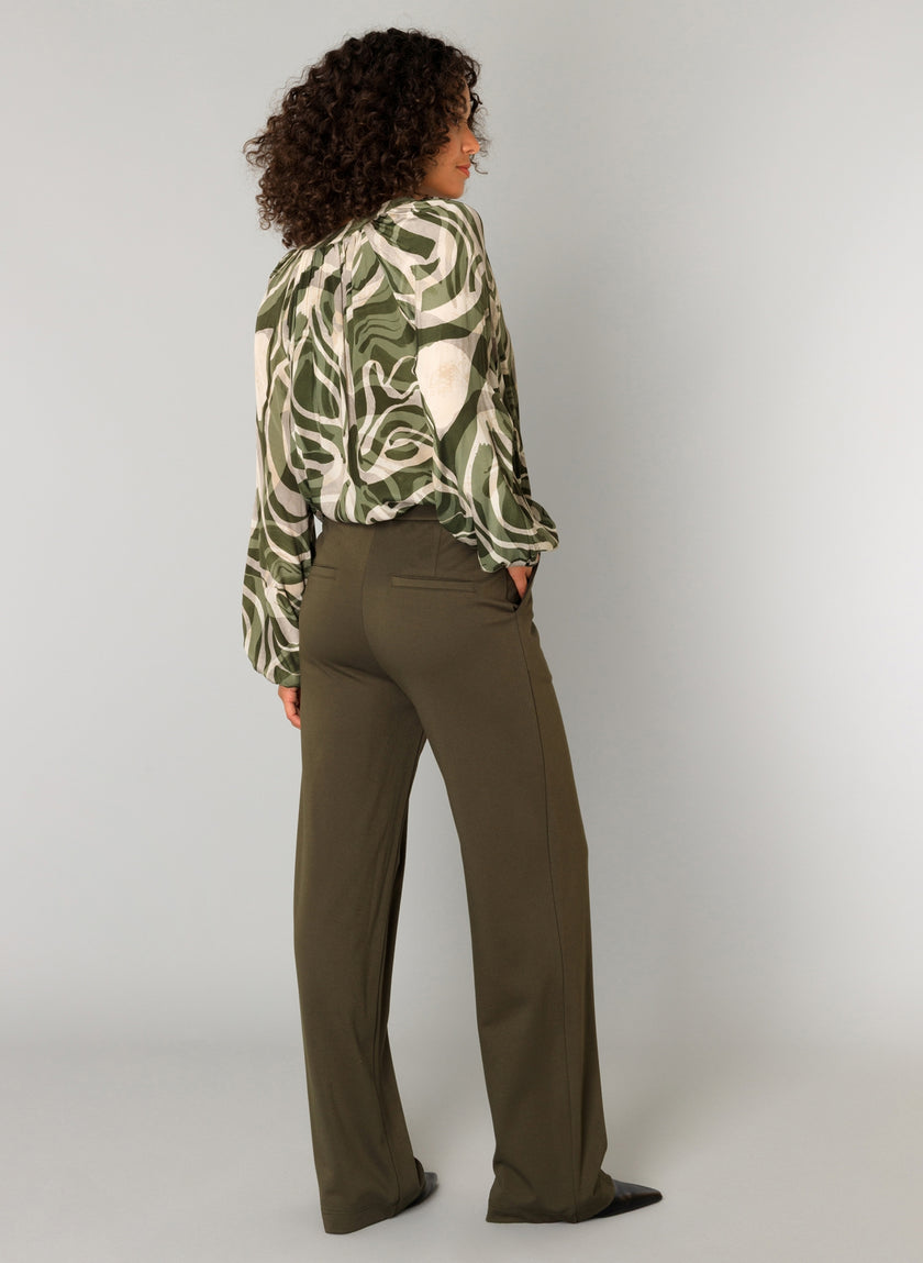 Een vrouw kijkt de andere kant op en draagt de Paloma Essential blouse in abstract groen en wit, gecombineerd met een olijfkleurige groene zachte pantalon met elastische tailleband en zwarte pumps, tegen een effen lichtgrijze achtergrond.