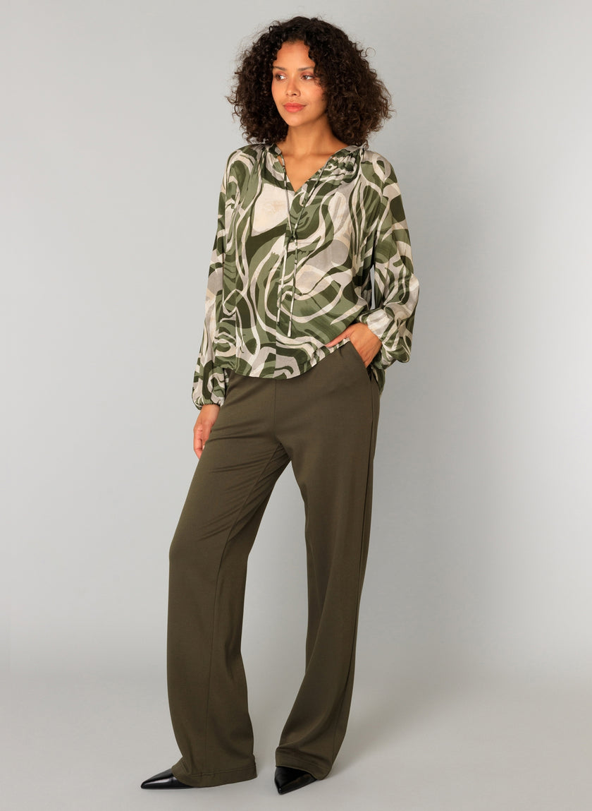 Een vrouw met krullend haar draagt de Paloma Essential blouse - verkrijgbaar in 4 kleuren - met een abstract groen en beige patroon, gecombineerd met een losse, zachte broek met elastische tailleband en zwarte schoenen met spitse tenen tegen een lichtgrijze achtergrond.