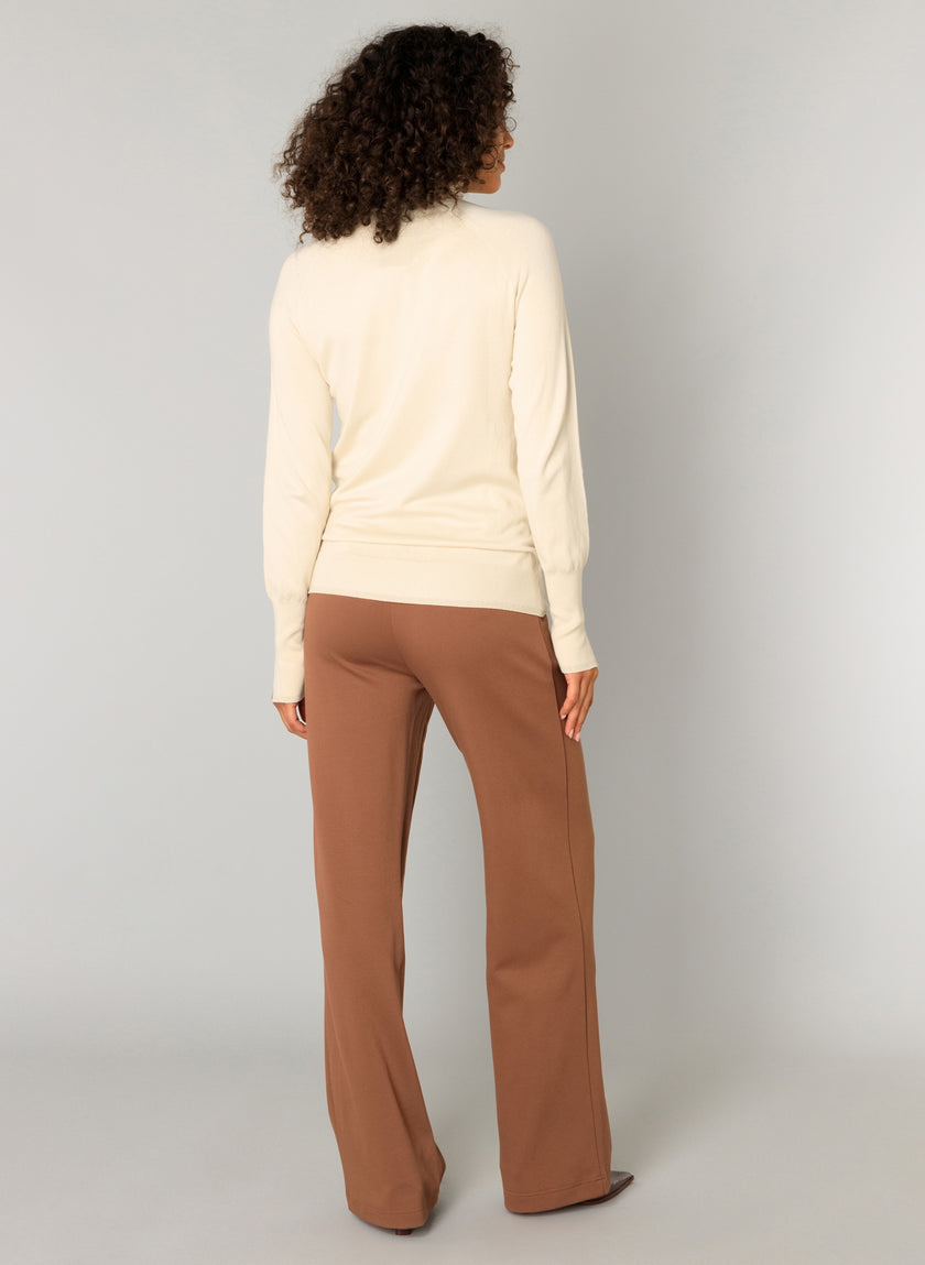 Een persoon met krullend haar kijkt weg, draagt een crèmekleurige top met lange mouwen en de Paloma Essential zachte pantalon met elastische tailleband, beschikbaar in 4 kleuren, tegen een effen lichte achtergrond.
