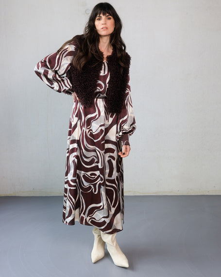 Een vrouw met lang donker haar draagt de Cristiana bordeaux print maxi-jurk in kastanjebruin en crème, gecombineerd met een vest met donkere textuur en witte hakken, staande tegen een effen lichtgekleurde muur.