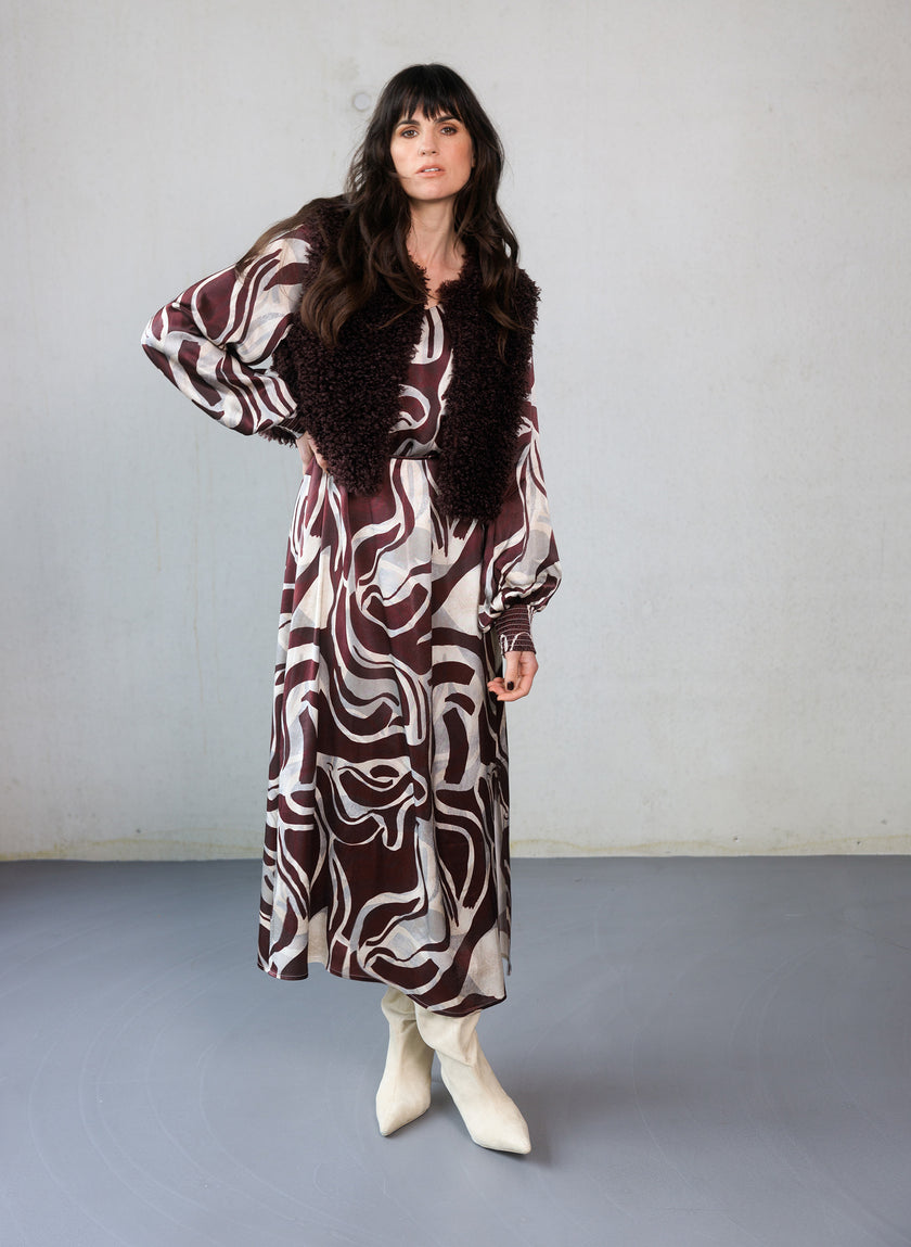 Een vrouw met lang donker haar draagt de Cristiana bordeaux print maxi-jurk in kastanjebruin en crème, gecombineerd met een vest met donkere textuur en witte hakken, staande tegen een effen lichtgekleurde muur.