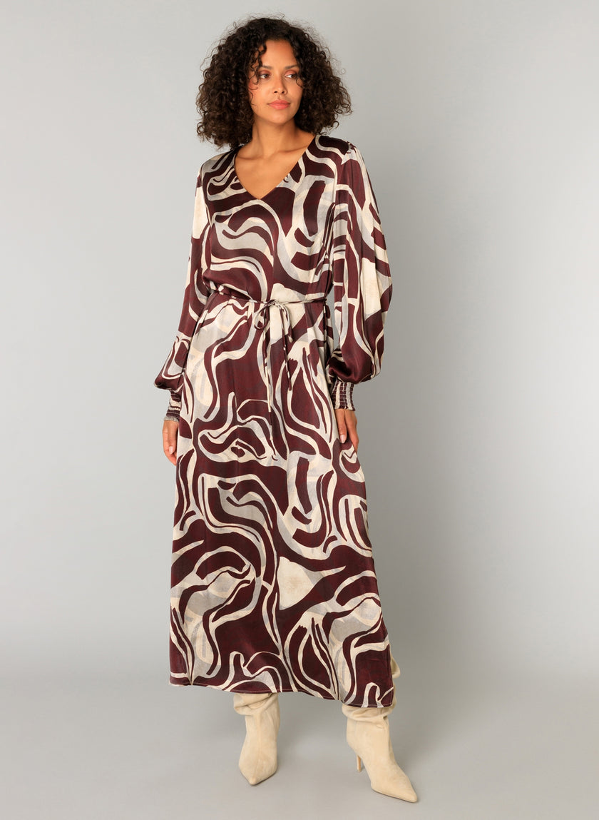 Een vrouw met krullend haar showt de Cristiana, een maxi-jurk met lange mouwen en riem met een opvallende abstracte print in bruin en crème, gecombineerd met laarzen met lichte hakken tegen een effen lichte achtergrond.