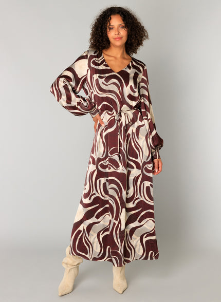 Een vrouw met krullend haar draagt de Cristiana bordeaux maxi-jurk met beige abstracte swirls, lange mouwen, een ceintuur in de taille en beige laarzen tegen een effen lichtgrijze achtergrond.