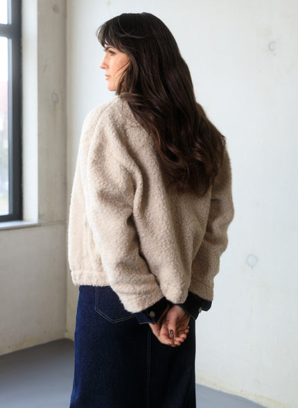 Een vrouw met lang bruin haar staat binnen bij een raam, met haar handen op haar rug geklemd. Ze draagt het Carmen-Maria jasje in beige en een donkere rok of broek - perfect voor het tussenseizoen.