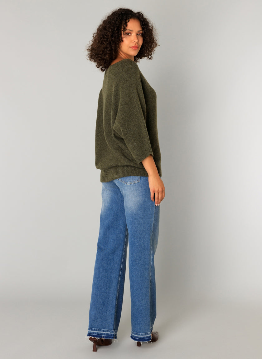 Een vrouw met krullend haar staat opzij en kijkt over haar schouder in een effen lichtgrijze omgeving. Ze draagt Cati - Legergroen, een losse trui in combinatie met een blauwe jeans met wijde pijpen en bruine schoenen met hak.