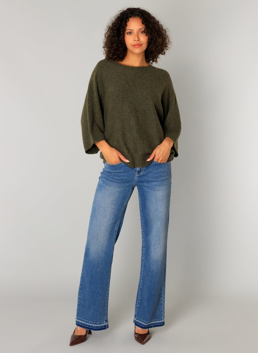 Een persoon met krullend haar draagt de oversized Cati - legergroene trui, gecombineerd met een blauwe spijkerbroek met wijde pijpen en bruine schoenen met spitse tenen, tegen een effen lichtgrijze achtergrond.