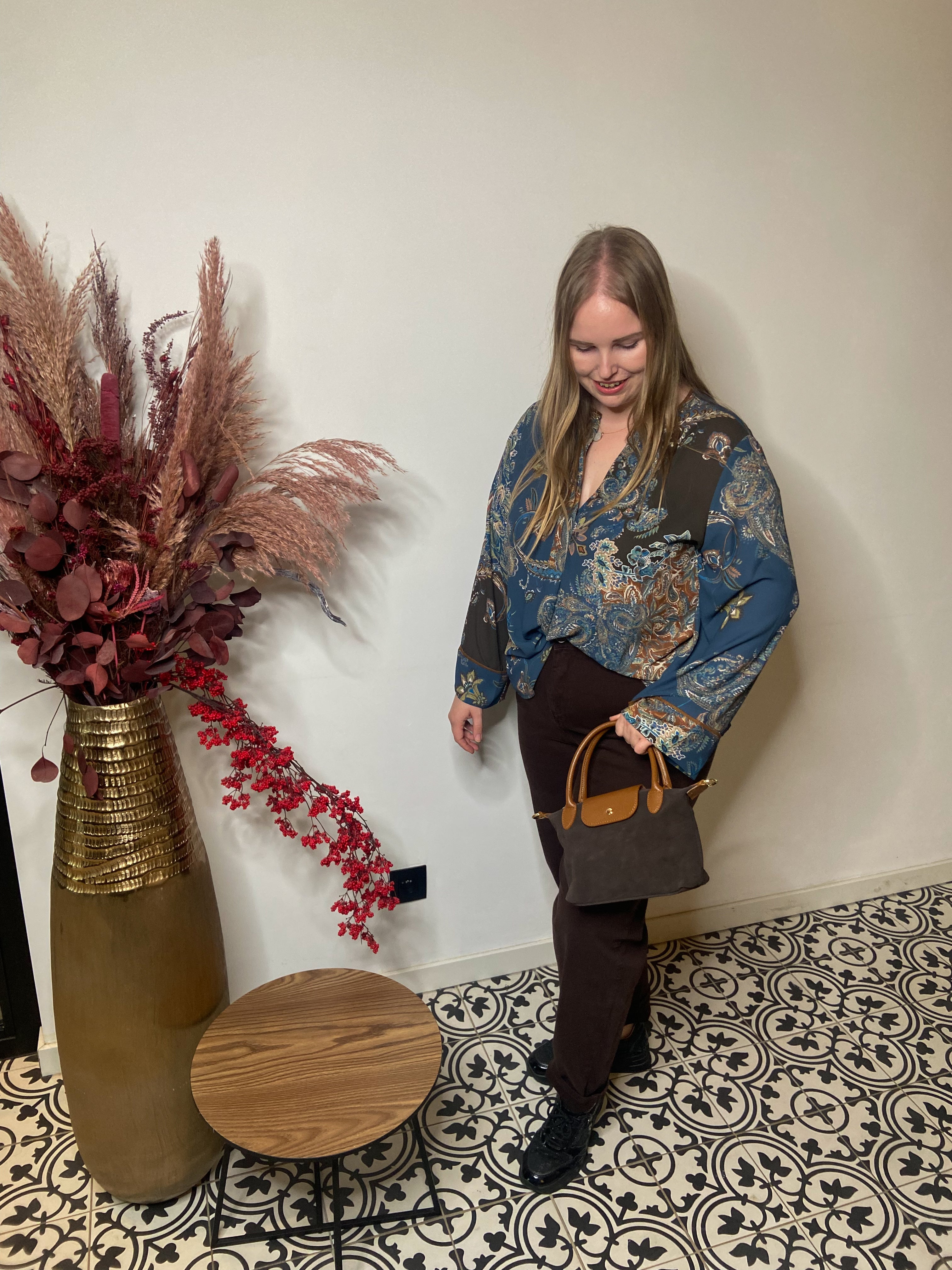 Een vrouw met lang blond haar draagt een Boheemse blouse - Mat Fashion, met wijde mouwen, gecombineerd met een donkere broek en een bruine handtas. Ze glimlacht binnenshuis op zwart-witte tegels en staat naast een vaas met gedroogde roze en rode bloemen.