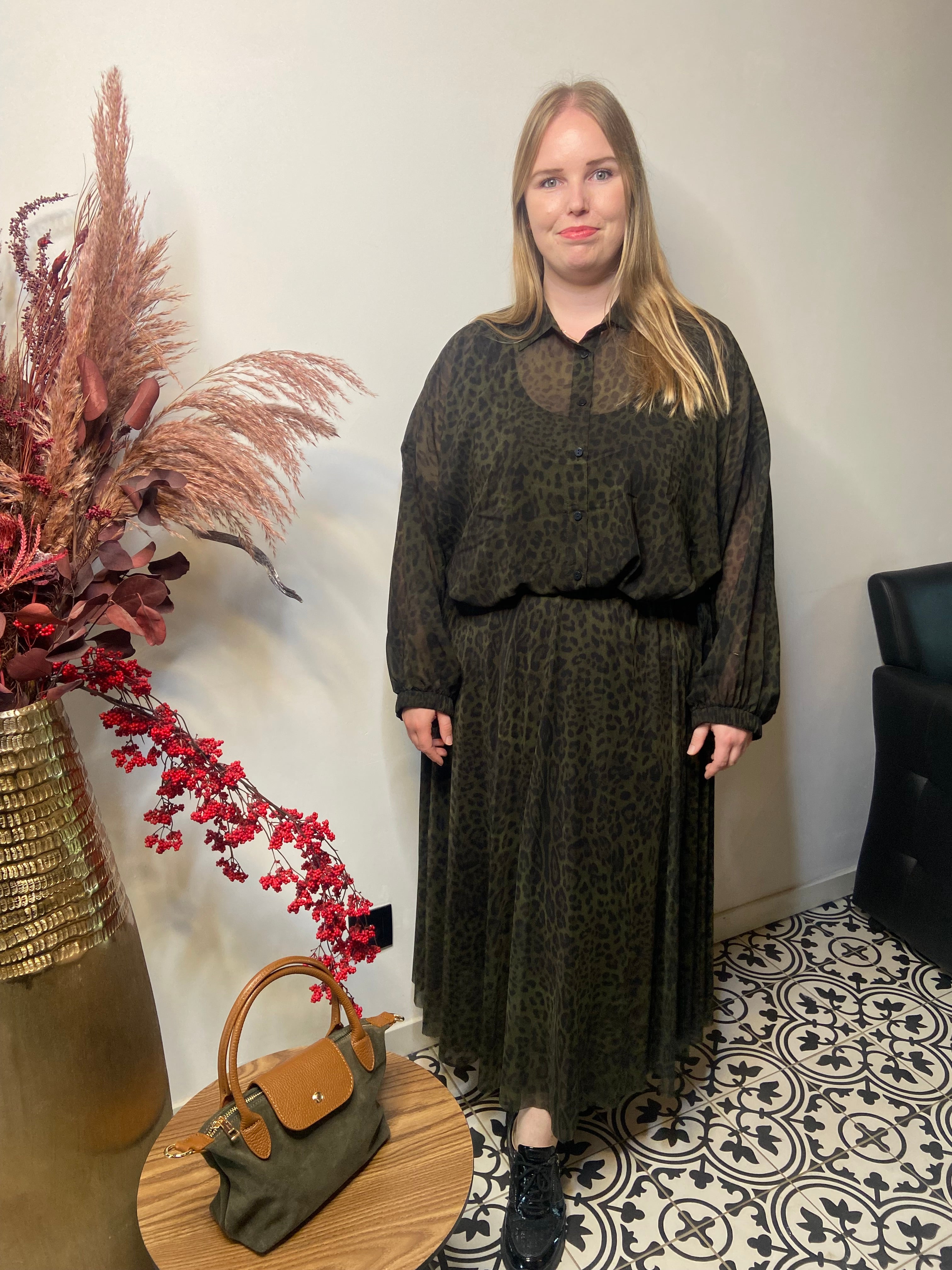 Een vrouw met lang blond haar staat binnen en draagt de Hemd met print - Mat Fashion en zwarte schoenen, naast een grote vaas met gedroogde planten en een tafeltje met een groene handtas op tegels met zwart en wit patroon.