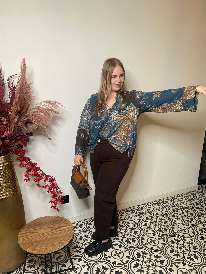 Een vrouw met lang blond haar glimlacht binnenshuis op tegels met een patroon, draagt een Boheemse blouse - Mat Mode met wijde mouwen, donkere broek, zwarte schoenen en houdt een bruine handtas vast. In de buurt staan decoratieve planten en een tafeltje.