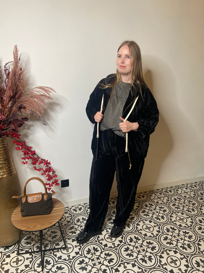 Een vrouw met lang blond haar staat binnen op gedessineerde tegels en draagt het Zwarte jas huispak - Mat Fashion over een grijs topje. Ze houdt de koorden van het jasje vast. Naast haar staat een klein tafeltje met een bruine handtas en gedroogde bloemen. 62043390.