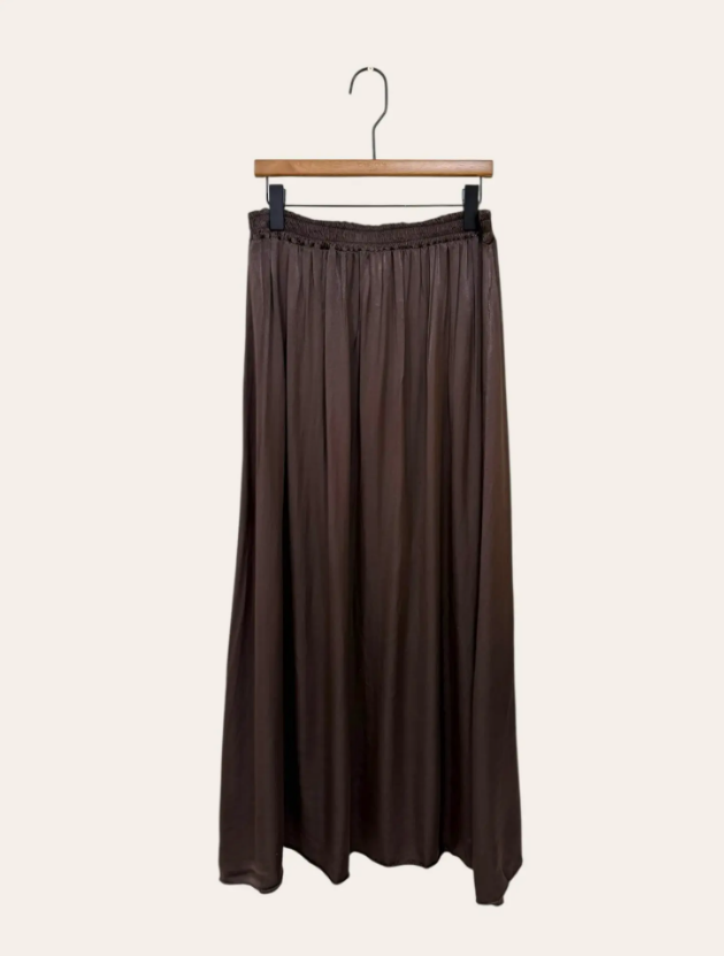 De Bruine rok is een lange, donkerbruine viscose rok met een vloeiend silhouet en een elastische tailleband, afgebeeld aan een houten hanger tegen een lichte achtergrond.