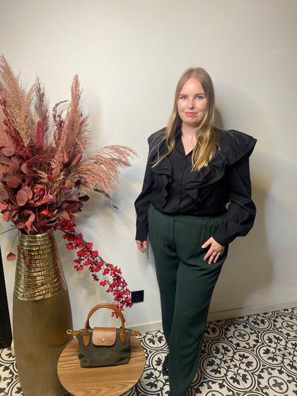 Een vrouw met lang blond haar staat binnen bij een hoge vaas met gedroogde bloemen, draagt een stijlvolle zwarte blouse met ruches op de schouders en een Groene broek van Mat Fashion, en staat naast een tafeltje met een groene handtas.