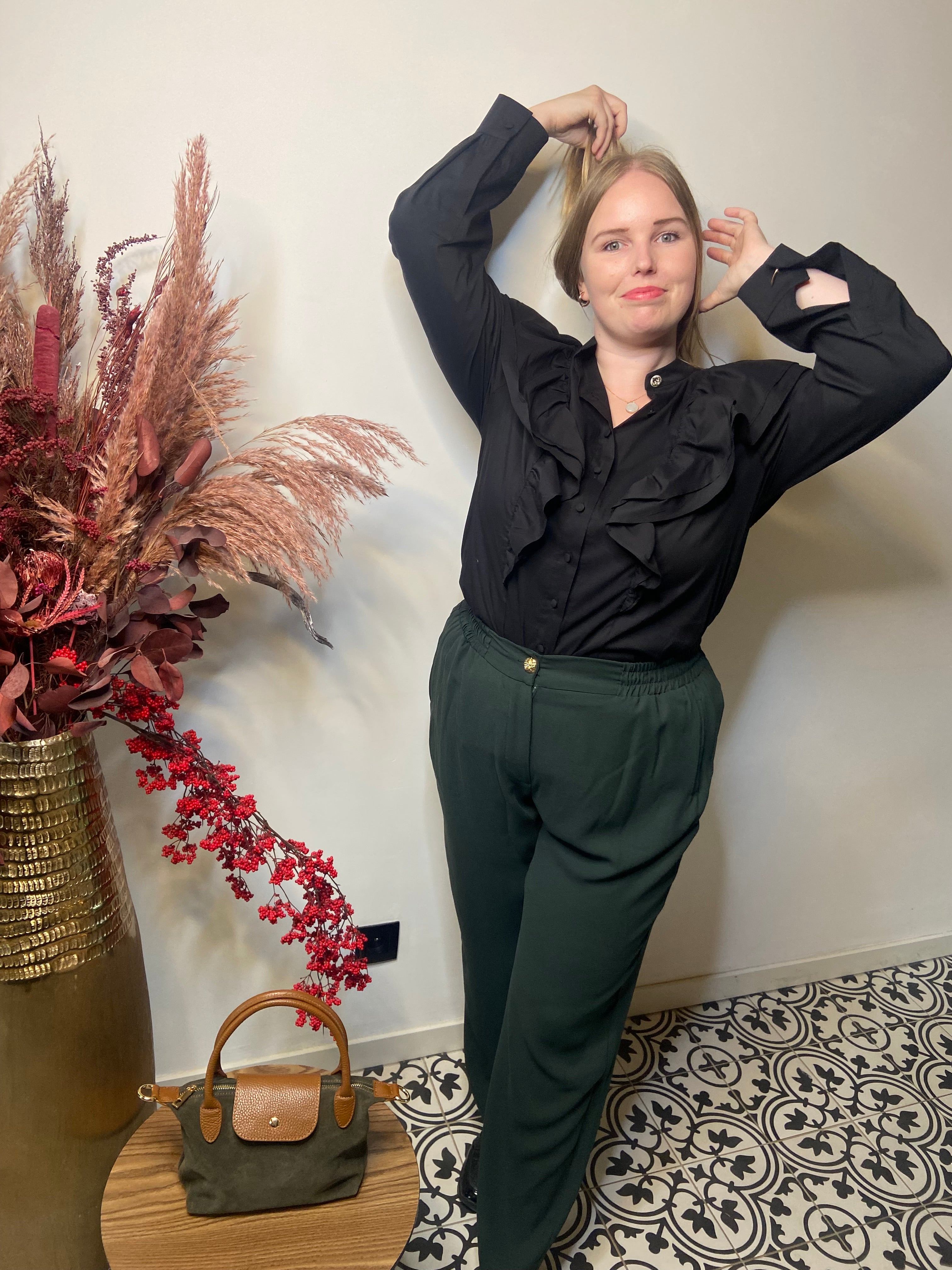 Een blonde vrouw poseert binnenshuis in een zwarte blouse en Groene broek - Mat Fashion. Ze staat bij een gouden vaas met gedroogde bloemen en een tafel met een groene handtas op een tegelvloer met patroon.
