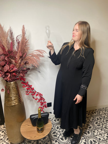 Een vrouw met het Zwart kleed van Mat Fashion staat binnen en glimlacht terwijl ze een lege champagneglas vasthoudt. Naast haar staat een hoge vaas met gedroogd roze en rood gebladerte, plus een tafeltje met een wijnfles en een zwarte tas.