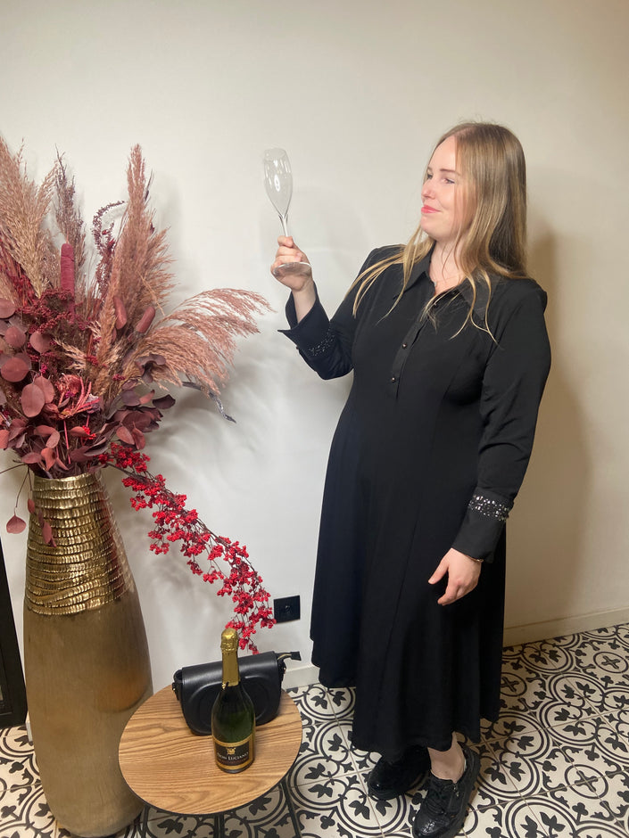 Een vrouw met het Zwart kleed van Mat Fashion staat binnen en glimlacht terwijl ze een lege champagneglas vasthoudt. Naast haar staat een hoge vaas met gedroogd roze en rood gebladerte, plus een tafeltje met een wijnfles en een zwarte tas.