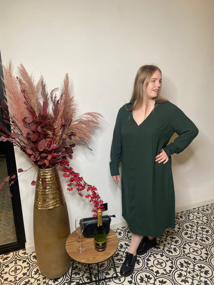 Een vrouw in een Groen kleed - Matmode staat op gedessineerde tegels naast een kleine ronde tafel met champagne, twee glazen en een zwarte tas. Een hoge vaas met gedroogde bloemen staat naast haar tegen een effen muur.