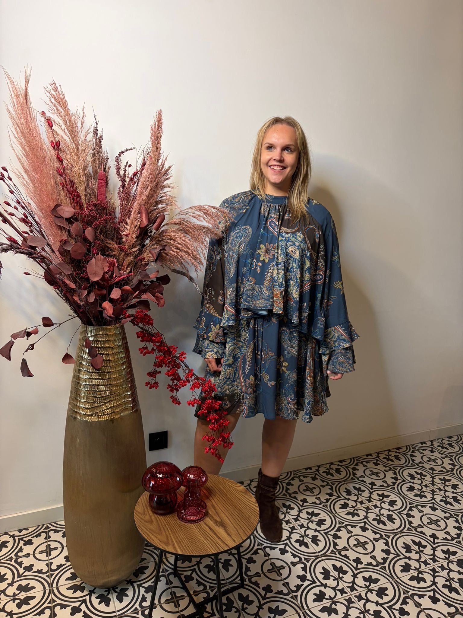 Een glimlachende vrouw in een blauw Boheems kleed indigo van Mat Fashion staat binnen naast een grote vaas met gedroogde planten en een kleine houten tafel met rood glazen decor op een zwart-wit geometrische tegelvloer.