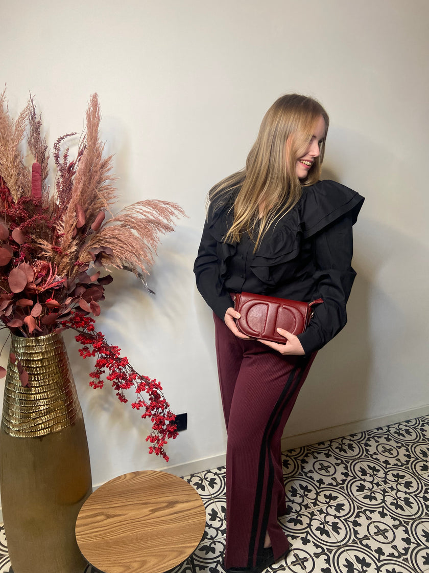 Een vrouw met lang blond haar staat binnen op een tegelvloer met patroon, ze draagt een Rode broek met strepen - Mat Mode en een zwart topje met ruches. Ze houdt een rode tas vast en glimlacht naast een ronde houten tafel en een hoge vaas met gedroogde planten.
