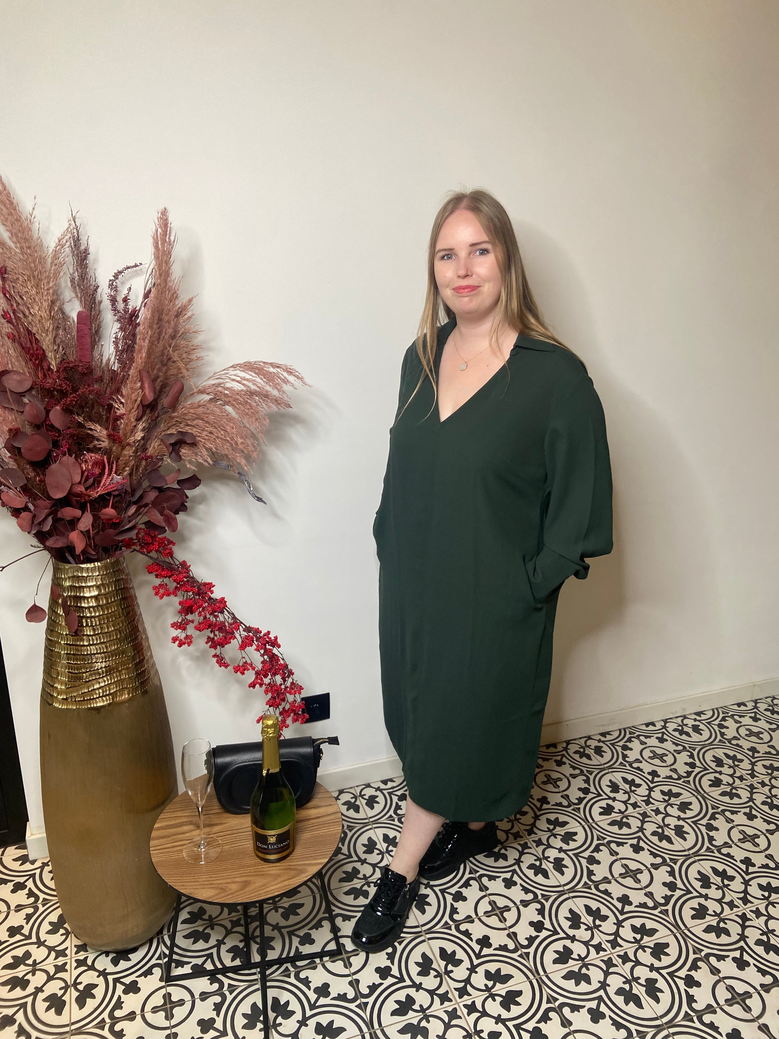Een vrouw met lang blond haar draagt het Groen kleed van Mat Fashion - een elegante, losse donkergroene jurk - terwijl ze naast een vaas met gedroogde bloemen en een gedekte tafel met champagne op een tegelvloer met patroon staat.
