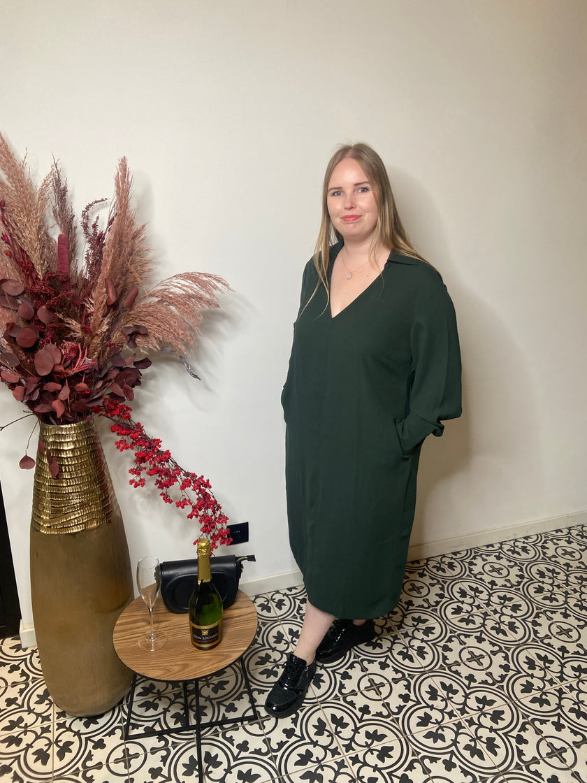 Een vrouw met lang blond haar draagt het Groen kleed van Mat Fashion - een elegante, losse donkergroene jurk - terwijl ze naast een vaas met gedroogde bloemen en een gedekte tafel met champagne op een tegelvloer met patroon staat.