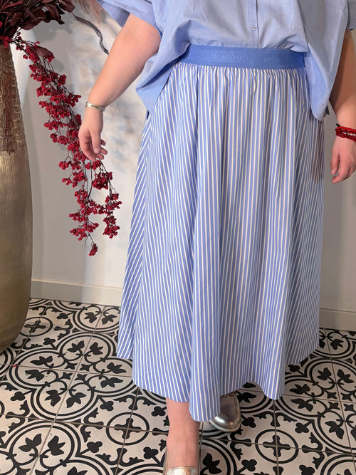 Dame draagt lichtblauwe gestreepte midi rok van Milano Italy met elastische tailleband, gecombineerd met een blauwe blouse.
