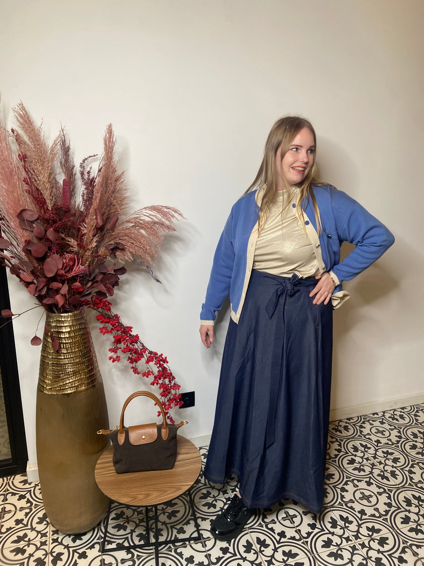 Een vrouw met een crèmekleurig topje, blauw jasje en de Denim rok van Mat Fashion poseert binnenshuis naast een grote vaas met gedroogde bloemen en een tafeltje met een bruine handtas, op tegels met een stijlvol patroon tegen een effen lichte muur.